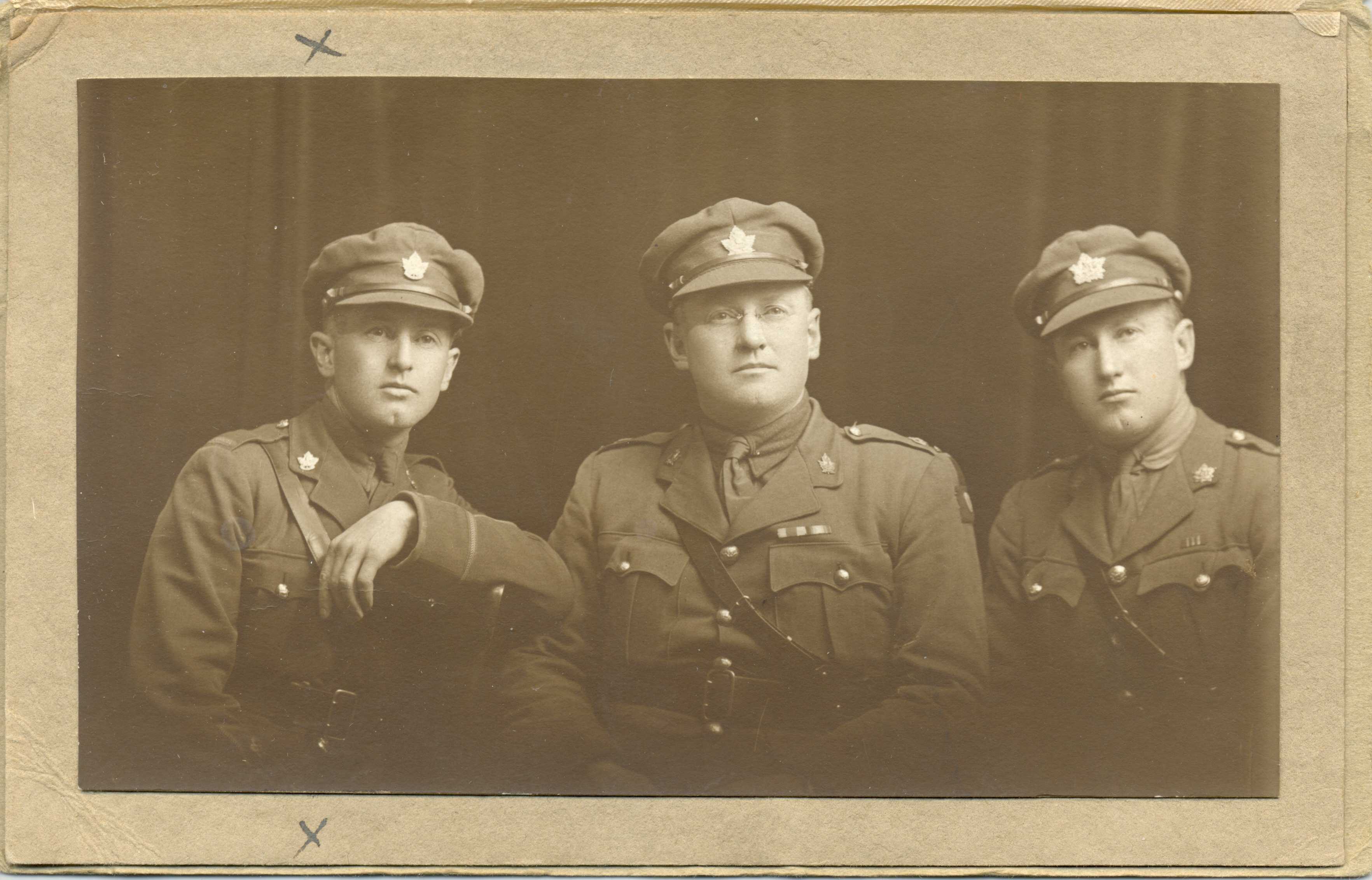 Sepia tone photo. Three men are posed in seated positions, facing the camera. They wear military dress, including caps.